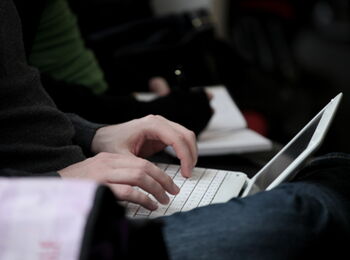 Two hands typing on a white notebook resting on the knees of the person writing.