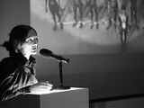 Black and white photograph of Vanessa Beecroft during a lecture. She stands at the lectern and speaks. In the background there is a screen showing naked people.