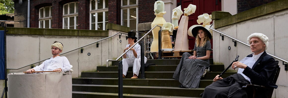 Four actors in historicising costumes sit on a staircase in front of a brick building, behind them stand various figures made of yellow and pink foam.