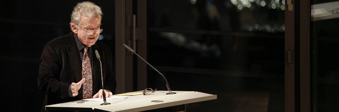Hans-Ulrich Gumbrecht stands at the lectern, brightly lit in a dark room, and speaks.