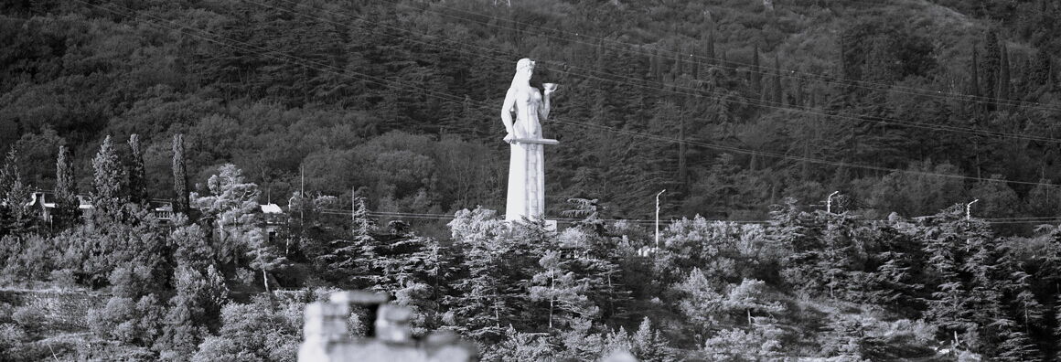 Black-and-white photograph of a large white female figure holding a bowl in her left hand and a sword in her right hand. She is surrounded by forest.
