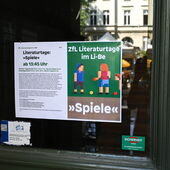 Announcement poster for the ZfL Literature Days at Li-Be, taped to a glass door reflecting the house across the street.