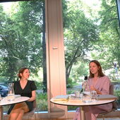 Marlen Hobrack und Kira Jürjens sitzen nebeneinander an weißen Bistrotischen und unterhalten sich. Durch die Fensterfront hinter ihnen sind Pflanzen, Bäume und eine Straße mit Autos zu sehen.