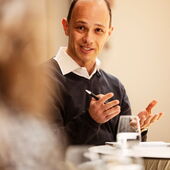 Alexander Libman sitzt an einem Bistrotisch und spricht. In der rechten Hand hält er einen schwarzen Kugelschreiber.