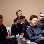 In drei grauen Stuhlreihen sitzen mehrere Zuhörer*innen. Einige schreiben mit, andere schauen konzentriert nach vorne.