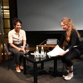 Elisa Aseva und Hanna Hamel sitzen auf einem Podium vor einer Leinwand. Hanna Hamel hält mehrere Blätter Papier in den Händen.