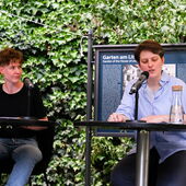 Christina Ernst and Anna Hetzer sit on a stage. Anna Hetzer reads from a book, Christina Ernst listens to her attentively.