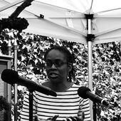 Eine Frau, Sharon Dodua Otoo, sitzt unter einem Sonnenzelt und gestikuliert. Vor ihr stehen Mikrofone.