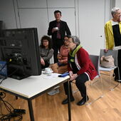 Three people are sitting on chairs and looking at a screen with interest, surrounded by three other people with coffee cups in their hands.