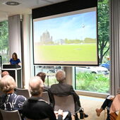 Uta Kornmeier steht am Rednerpult. Neben ihr auf der Leinwand ist eine grüne Wiese mit dem Fernsehturm und dem Berliner Dom im Hintergrund zu sehen.
