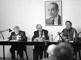 Black and white photograph of xxx, Carlo Ginzburg and Robert Stockhammer. The three sit on a podium in front of a crowd. A picture of Erich Auerbach hangs in the background.