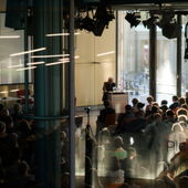 Wide shot of a crowded hall, at the front stands Homi Bhabha behind a lectern and gives a speech