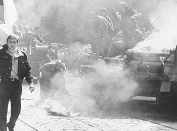 Schwarzweißfoto einer Straßendemonstration mit Rauch, einem brennenden Panzer und Menschen, die Flaggen tragen.