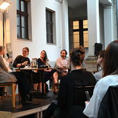 The speakers sit on a podium in front of an audience in a courtyard. Martina Stemberger speaks into a microphone.