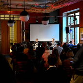 In the background Elias Hirschl reads from his book, Hanna Hamel listens. An audience sits in the foreground.