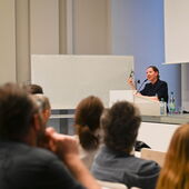 Maggie Nelson stands at the lectern and speaks to the audience. In her right hand she holds her glasses.