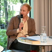 Patrick Hohlweck sitzt an einem weißen Bistrotisch und spricht ins Mikrofon.