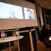 Ulfa von den Steinen und Sandra Folie sitzen halb zur Leinwand gewandt neben dem Rednerpult. Auf der Leinwand sind die Zoom-Kacheln von Jürg Schneider und Philipp Khabo Koepsell zu sehen.