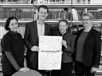 Artemis Alexiadou, André Lottmann, Eva Geulen, and Ulrike Freitag stand in front of a bookshelf. Lottmann and Geulen are holding a poster with the hashtag #Zusammenland.