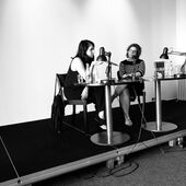 Black and white photograph of Fatma Aydemir and Sonja Longolius sitting on a podium. Fatma Aydemir gestures with her right hand, Sonja Longolius looks at her.