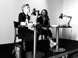 Black and white photograph of Esther Kinsky and Mona Körte, the two are sitting on a podium. Esther Kinsky speaks to the crowd, Mona Körte looks at her.