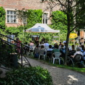 Im Garten des Literaturhauses sitzen circa 20 Personen auf Gartenstühlen, sie tragen Headsets und schauen auf ein Gartenzelt mit Bühne, auf der Janika Gelinek mit Heike Geißler spricht.