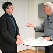 Ernst Müller is standing in front of the screen and speaking; Dirk Naguschewski can be seen in the background, looking in his direction.