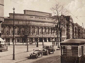 Black-and-white photo: A historic street scene featuring a curved corner building with the word “Atrium” in the center, surrounded by shops, a streetcar, and passersby.