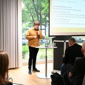 Sebastian Kirsch steht vor dem Publikum und spricht ins Mikrofon. Neben ihm auf der Leinwand sind die Namen der nächsten Vortragenden zu sehen.