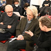 Three people are sitting next to each other on black and red chairs. The person on the right gestures and talks to the other two, who listen to them.