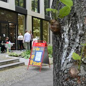 Rechts ein Baumstamm, links im Hintergrund ein Aufsteller mit Veranstaltungsplakat und mehrere Personen, die vor dem Eingang zum Veranstaltungsraum stehen und sitzen.