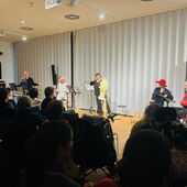 Five actors and musicians sit and stand between music stands, with well-filled rows of audience members in front of them.