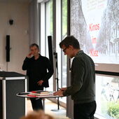 Dirk Naguschewski stands at a bistro table with books and speaks, Christoph Hesse can be seen in the background at the lectern. “Kinos im Kiez” is written on a screen behind them.