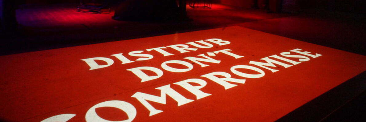 In einem dunklen Raum ist auf rotem Grund in weißen Lettern der Schriftzug "Distrub don't compromise" zu lesen.