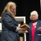 Eine Person übergibt einer anderen zwei alte Bücher, im Hintergrund steht ein Bücherregal.