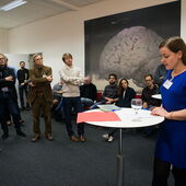 Stefanie Burkhardt steht an einem Tisch und liest vor, eine Gruppe Zuschauer*innen hört ihr zu