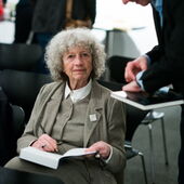 Ulrike Ottinger sits on a chair with a book in her lap and writes something in it, next to her stands a man