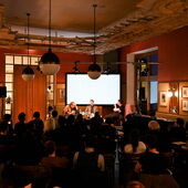 You can see a large room filled with warm light. The audience is seated in the first half, with three people on a podium at the end, with a bright screen behind them.