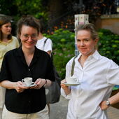 Hanna Hamel und Katrin Trüstedt stehen gemeinsam im Garten des Literaturhauses und halten Kaffeetassen in den Händen, im Hintergrund sind zwei Postkartenständer zu sehen.