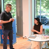 Georg M. Oswald steht vor dem Rednerpult und unterhält sich mit Lin Hierse, die an einem weißen Bistrotisch sitzt.