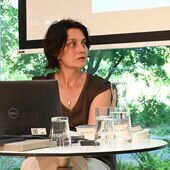 Katja Petrowskaja sitzt an einem weißen Bistrotisch, auf dem ein Laptop steht, und schaut nach rechts.