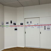 Auf einer weißen Wand und Schiebetüren ist ein horizontaler blau-grün-roter Klebestreifen angebracht, um den herum Zettel mit verschiedenen Jahreszahlen, Bildern und kurzen Texten gruppiert sind.