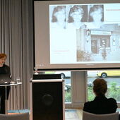 Nina Weller stands at a bistro table with a laptop on it next to the lectern and speaks to the audience. On the screen to her right is a picture of Wanda Vrubliauskaite and one of the “Kleine Philharmonie”.