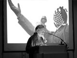Black and white photograph of W. J. T. Mitchell. He is standing at a lectern giving a lecture. In the background is a large screen showing two men.