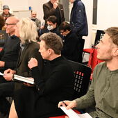 Four people sit on black and red chairs next to each other and look to the right. In the background you can see more people sitting on chairs.