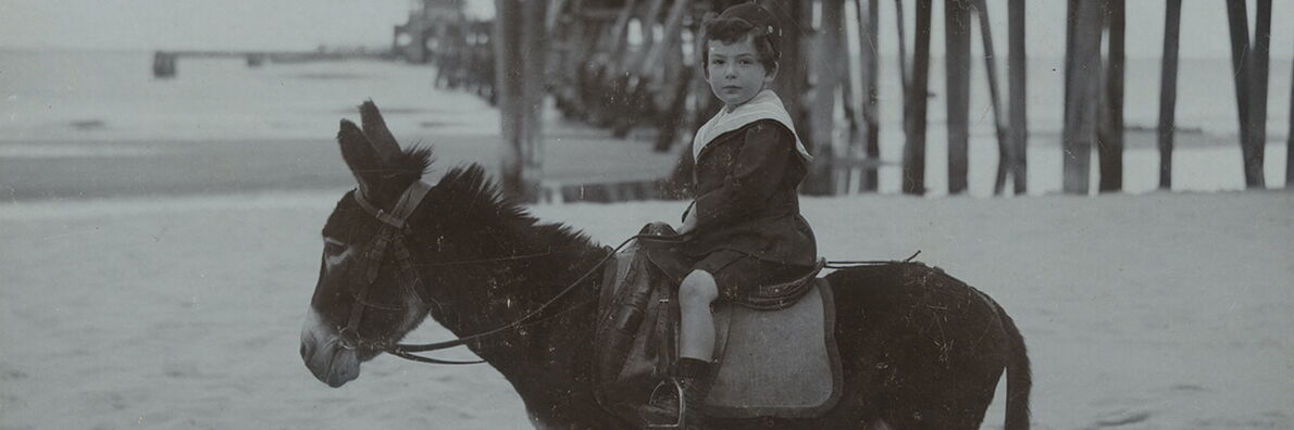 A black-and-white photo of a young boy wearing a cap and shorts, sitting on a donkey on the beach. A pier can be seen behind him.