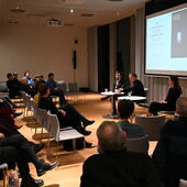 In einem großen Raum mit Parkettboden sitzen ca. 20 Personen im Publikum, vorne sitzen drei Personen vor einer Leinwand, auf der das Buch "The Conspiracy of the Carpenters" zu sehen ist.