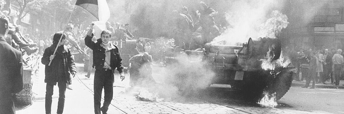 Black-and-white photo of a street protest with smoke, a burning tank, and people carrying flags.