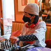 Die DJ Mo Chan / DJ Kohlrabi  steht am Mischpult. Sie trägt eine Mütze und eine FFP2-Maske.