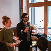 Hanna Hamel und Andreas Scherffig stehen vor einem Podium. Scherffig hält ein Mikrofon in der Hand und spricht.